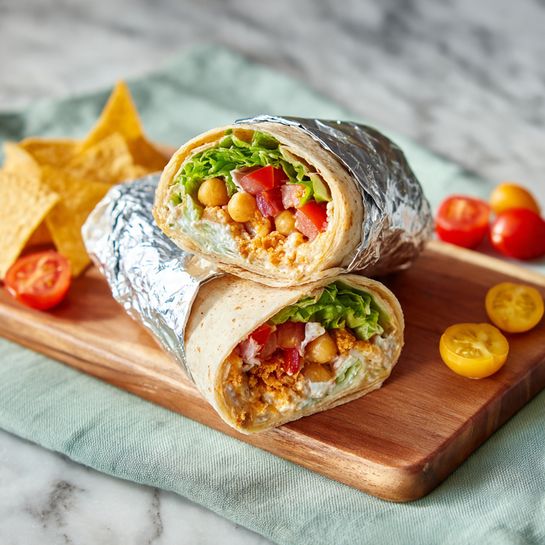 A close-up of a wrap sandwich cut in half and wrapped in foil, placed on a small rectangular wooden board laid on a light green cloth. The wrap shows visible layers inside: a soft light beige tortilla, filled with golden brown chickpeas, bright green lettuce, red tomato slices, and bits of white sauce or cream. The wrap halves are stacked slightly, with the top half leaning against the other. Around the board, there are a few yellow triangle chips and halved cherry tomatoes. The scene is set on a white marbled surface. photo taken with an iphone --ar 4:5 --v 7
