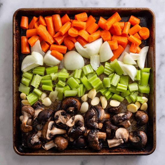 The image shows a textured baking tray filled with colorful chopped vegetables and mushrooms. There are three layers from top to bottom: the top layer has thick pieces of bright orange carrots and quartered white onions with their smooth outer skin visible; the middle layer consists of chopped green celery sticks with a fresh, crisp texture and whole peeled garlic cloves scattered around; the bottom layer features large pieces of dark brown mushrooms with white edges and black gills underneath. The vegetables look fresh and lightly coated with oil, all arranged closely but not overlapping on the baking tray, set on a white marbled surface. photo taken with an iphone --ar 4:5 --v 7