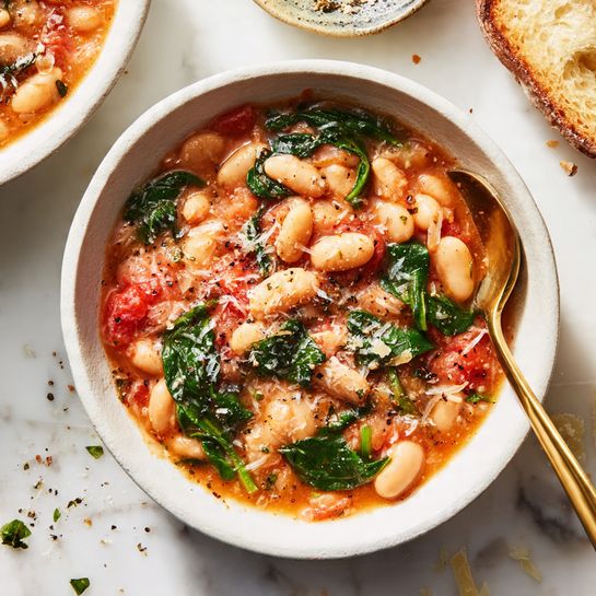 A white pot is filled with a thick stew showing three main layers: at the bottom, a rich reddish-brown sauce with visible small chunks of soft tomatoes; the middle layer reveals light orange carrot slices and creamy white beans mixed in, adding texture; the top layer has dark green leafy vegetables scattered throughout, giving a fresh contrast to the warm colors. The inside edges of the pot are coated slightly with the sauce, and the pot is set on a white marbled surface photo taken with an iphone --ar 4:5 --v 7