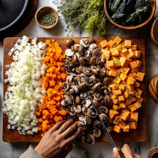 A wooden cutting board filled with neatly chopped vegetables, arranged in groups: white onion pieces on the left, bright orange carrot slices above them, and cubed sweet potatoes on the right. A woman's hand is holding down sliced brown mushrooms in the center, while the other woman's hand is slicing more mushrooms with a knife. The background shows fresh green herbs at the top and a white marbled cutting board with a knife and dark leafy greens on the upper right. A black cast iron pot lid is partially visible in the top left corner. The scene is lit warmly and looks ready for cooking photo taken with an iphone --ar 4:5 --v 7