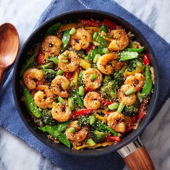 A black pan filled with a colorful shrimp stir-fry, showing three main layers: the bottom layer has light brown cooked garlic bits and oil, the middle layer includes bright green broccoli florets and sugar snap peas mixed with red and yellow bell pepper chunks, and the top layer is covered with orange shrimp curled up, sprinkled with white and black sesame seeds and scattered sliced green onions. The pan rests on blue cloth over a white marbled surface with a wooden spoon near the bottom left corner. Photo taken with an iphone --ar 4:5 --v 7