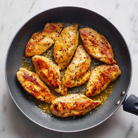 A gray pan holds eight golden brown pieces of cooked chicken tenders arranged in a loose circle with some oil glistening underneath, creating a shiny texture. The tenders have a slightly crispy, textured surface with some browning on top, and steam rises gently from the pan, hinting they are hot. The pan sits on a white marbled surface that adds subtle light reflections to the scene. photo taken with an iphone --ar 4:5 --v 7