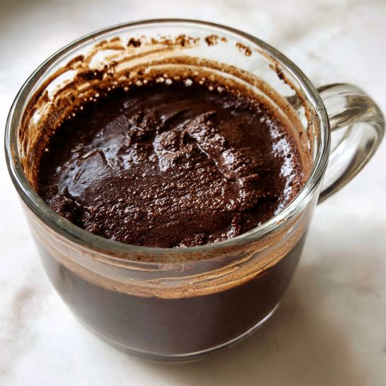 A clear glass mug filled with a thick, dark brown chocolate mixture that looks dense and slightly glossy. The mixture has textured swirls and small lumps, showing rich and moist chocolate inside the mug. The mug is placed on a white marbled surface, and some chocolate smudges are visible around the base of the mug. Photo taken with an iphone --ar 4:5 --v 7
