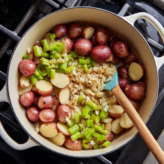 Inside a white pot on a stove with a black inside, there are three main layers of ingredients being cooked and stirred with a wooden spoon that has a light blue silicone head. The bottom layer is small white and light brown diced pieces that look soft and cooked. On top of that, there are bright green celery pieces cut into curved slices. Spread through the dish, there are many halved small red potatoes with smooth skin and pale yellow centers. The white pot handles shine in the light, and the scene shows part of the stove’s black cast iron grates under the pot. Photo taken with an iphone --ar 4:5 --v 7