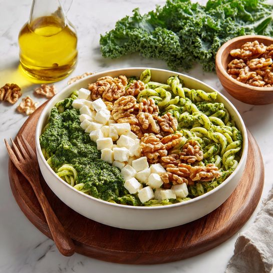 A large white shallow bowl filled with three layers: the bottom layer of short twisted pasta covered in vibrant green pesto sauce; the middle layer of small white cheese cubes scattered evenly throughout; and the top layer of medium brown walnut pieces sprinkled all over. The bowl is placed on a round wooden board with a wooden fork resting beside it. In the background, a glass bottle with yellow oil and a wooden bowl with more walnuts sit on a white marbled surface along with fresh green kale leaves. Photo taken with an iphone --ar 4:5 --v 7