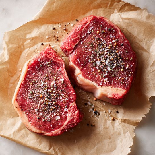 Two thick raw steaks rest on light brown parchment paper that covers a white marbled surface. Each steak has a pinkish-red center with a light layer of fat along one edge. The steaks are sprinkled with coarse salt and cracked black pepper, creating a textured contrast on the meat's surface. The close-up view shows the detail of the seasoning and the freshness of the steaks. Photo taken with an iphone --ar 4:5 --v 7