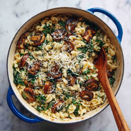 The image shows a white pot filled with a pasta dish. The pasta is short and twisted, mixed with browned mushroom slices and wilted dark green leafy vegetables. Shredded white cheese is scattered on top, melting slightly. A wooden spoon with some pasta and mushroom slices is resting inside the pot on the right side. The pot has blue handles, and it is placed on a white marbled surface. The whole dish looks warm and rustic. photo taken with an iphone --ar 4:5 --v 7