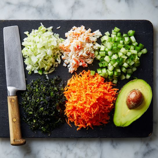 The image shows a black cutting board on a white marbled surface with five piles of chopped or shredded ingredients arranged in a loose circle. From the top left, there is a pile of small white and orange pieces resembling imitation crab meat, next to a pile of small green cucumber cubes. Below the cucumber is a bright orange pile of thin shredded carrots. To the left of the carrots, there is a dark green pile of chopped seaweed pieces. Below the imitation crab meat and seaweed, there is half an avocado; one half is whole and hollowed out, while the other half is chopped into cubes. A large knife with a silver blade rests on the left side of the board. Photo taken with an iphone --ar 4:5 --v 7