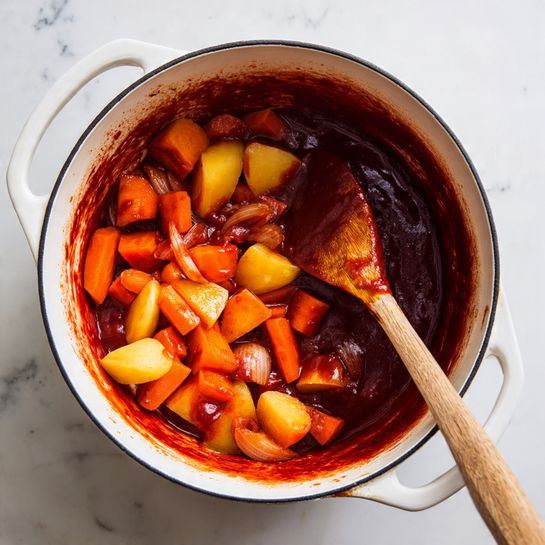Inside a white pot with a dark rim, there is a mix of cooked vegetables including orange carrot slices, golden yellow potato chunks, and small pieces of translucent white onion. The vegetables are coated in a deep red sauce that clings to the sides of the pot, showing a thick and rich texture. A wooden spoon with sauce residue is resting inside the pot, angled to the right. The pot sits on a white marbled surface. Photo taken with an iphone --ar 4:5 --v 7