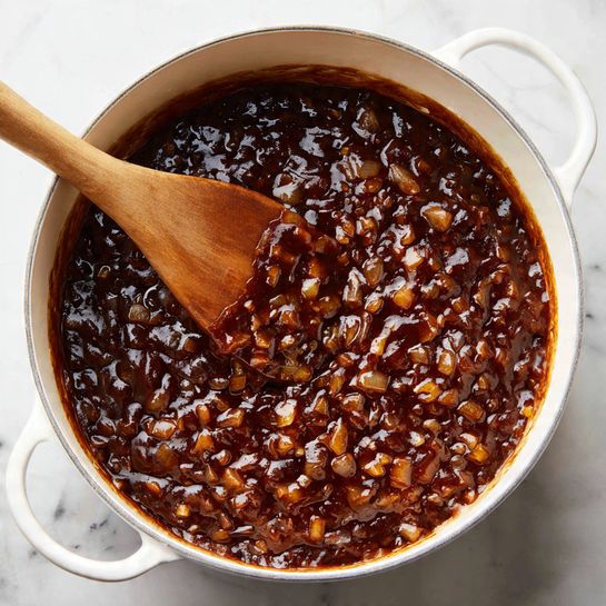 A close-up view of a white pot filled with a dark brown, glossy sauce mixed with translucent, small chopped onions all over. A wooden spoon is soaking in the sauce, showing the mixture's thick, shiny texture as it stirs the layers together. The pot is sitting on a white marbled surface, and the handle of the pot is visible on the right side. The sauce looks rich and full of flavor. photo taken with an iphone --ar 4:5 --v 7