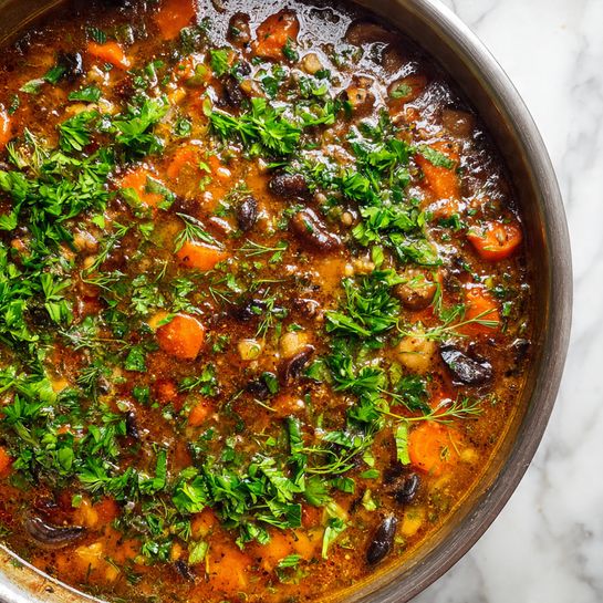 A close-up view of a pan filled with a thick stew showing three main layers: at the bottom, a rich brown broth with a slightly oily texture; in the middle, orange carrot chunks and small dark brown mushroom pieces scattered evenly; on top, finely chopped fresh green herbs spread all over, adding a bright contrast. The pan's edge is silver and the background is a white marbled texture. photo taken with an iphone --ar 4:5 --v 7