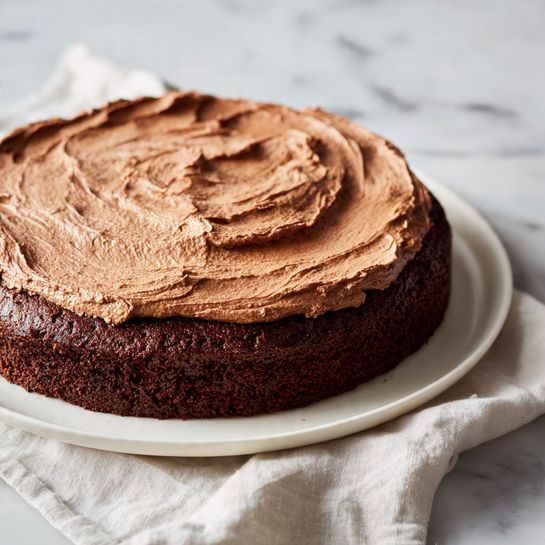 A single-layer dark chocolate cake is shown placed on a white plate, sitting on a white marbled surface with a soft white cloth nearby. The cake has a thick, smooth layer of light brown chocolate frosting evenly spread across the top, with visible creamy swirls and texture created by a spreading tool. The focus is on the top surface and edges of the cake, showing the contrast between the dark cake base and the lighter frosting. photo taken with an iphone --ar 4:5 --v 7
