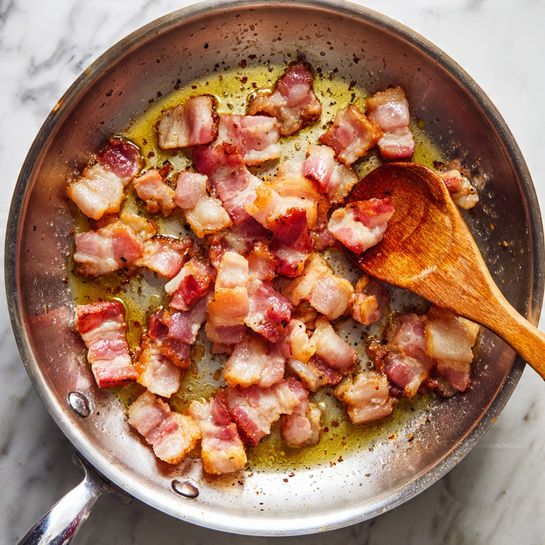 A close-up of small pieces of bacon frying in a shiny steel pan on a stove. The bacon is in the center of the pan, showing a mix of pink and golden brown colors, with oil around it spreading shiny spots on the pan’s surface. A wooden spoon is stirring the bacon from the side while the pan handle is barely visible on the upper left. The background is a white marbled texture. photo taken with an iphone --ar 4:5 --v 7