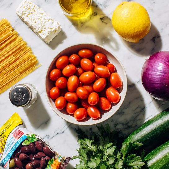 The image shows a white bowl filled with shiny, red grape tomatoes placed near the center on a white marbled surface. Surrounding the bowl, there are several fresh ingredients laid out neatly: a block of creamy white feta cheese on the top left, a bright yellow lemon next to a purple-red onion on the top right, a long dark green cucumber on the right edge, and fresh green herbs including parsley and basil arranged nearby. On the bottom left, a clear glass salt grinder stands next to an unopened bag of uncooked golden orzo pasta with a colorful label, and a jar of dark purple kalamata olives with a white label sits between the herbs and pasta. A clear bottle filled with golden olive oil is located near the top left corner. The whole scene is brightly lit, showing the vivid textures and colors of each item in sharp detail, with a clean and fresh presentation photo taken with an iphone --ar 4:5 --v 7
