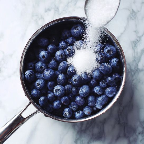 A silver cooking pot filled with a large number of fresh blueberries at the bottom. White granulated sugar is being poured over the blueberries from above, partially covering them. The pot sits on a white marbled surface, and the overall scene is bright and clear. Photo taken with an iphone --ar 4:5 --v 7