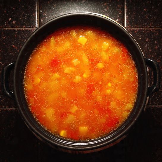 A round black pot filled with orange-red soup that has small chunks of yellow and lighter orange vegetables scattered inside, placed on a dark tiled surface with a folded cloth underneath the pot's handle, the soup’s surface shows some small oil bubbles and a smooth texture along the edges, photo taken with an iphone --ar 4:5 --v 7