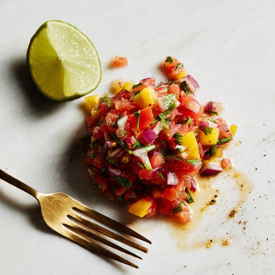 This image shows a pile of colorful salsa on a white marbled surface. The salsa has red pieces, yellow chunks, and green herbs mixed together, creating a fresh look with small bits of red onion. Two gold forks are placed on either side, one holding part of the salsa. A lime half sits on the left side, showing its bright green inside. The salsa looks moist with some juice around it on the white marble. photo taken with an iphone --ar 4:5 --v 7
