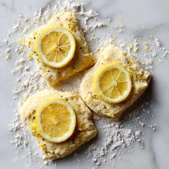 The image shows three square pieces of dough placed on a white marbled surface dusted with flour. Each dough piece is pale yellow with a slightly rough and uneven texture, sprinkled lightly with bits of zest or small particles. On top of each square dough piece lies a round, thin slice of lemon, showing bright yellow color with visible segments and a slightly translucent look. The flour is scattered unevenly around the dough pieces, creating a soft contrast with the smooth white marbled background. photo taken with an iphone --ar 4:5 --v 7