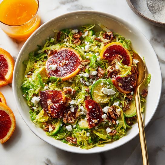 A large white bowl with a mix of chopped green Brussels sprouts forming the base layer, topped with scattered slices of deep red blood oranges that show a juicy texture. Crumbled soft white cheese is sprinkled evenly on the top layer, along with small pieces of brown nuts adding crunch. Light brown toasted crumbs are spread gently over the dish. The bowl is placed on a white marbled surface with some blood orange slices and a small glass bowl with orange dressing and a spoon visible nearby. Photo taken with an iphone --ar 4:5 --v 7