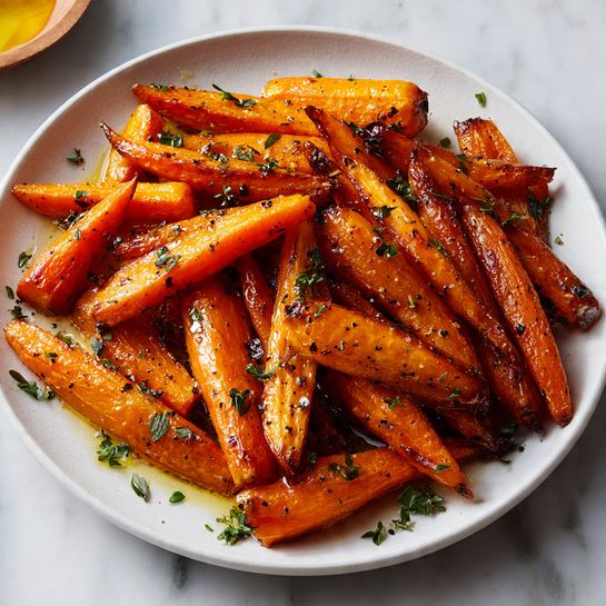 A white plate holds a pile of roasted carrots, each piece cut into thick sticks with a rich orange color and slightly charred edges. The carrots have a glossy, caramelized texture with a few scattered black pepper specks and small green herb bits sprinkled evenly on top. The plate is placed on a white marbled surface with some herbs and a wooden bowl with honey partially visible at the edges. The overall look is warm and inviting, with the carrots arranged naturally but densely on the plate. photo taken with an iphone --ar 4:5 --v 7