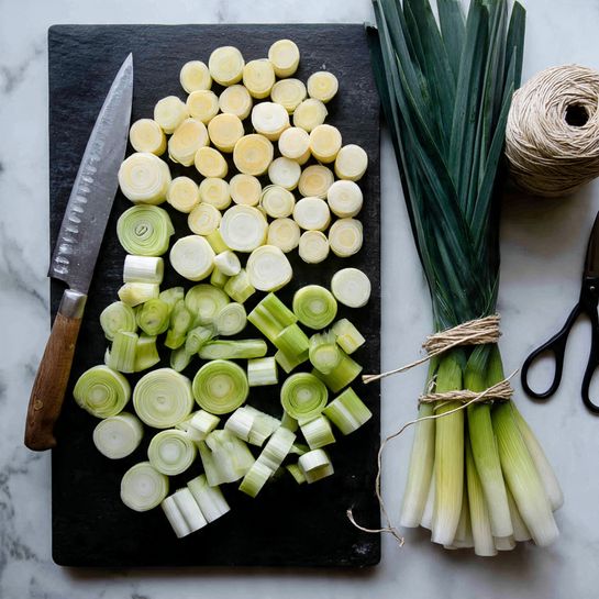 The image shows two layers. The top layer has many sliced rounds of light green and white leek pieces spread out on a dark cutting board with a large silver knife on the left side. The leek slices have a mix of pale yellow, white, and green colors with visible ring patterns. The bottom layer shows two bundles of long dark green leek leaves tied with string, placed on a dark cutting board. To the right of the bundles, there is a spool of beige string and a pair of black scissors lying flat. The background is a white marbled surface photo taken with an iphone --ar 4:5 --v 7