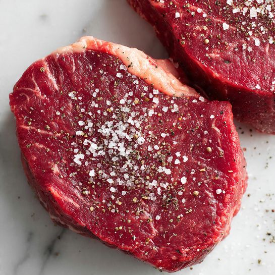 Two round raw steaks are shown on a white marbled surface. Each steak has a thick layer of deep red meat with light marbling of fat. On top, there is a generous sprinkle of coarse white salt and cracked black pepper, creating a textured look with white and dark specks. The close-up view shows the steaks slightly overlapping with one near the top right and the other partially visible at the bottom left. Photo taken with an iphone --ar 4:5 --v 7