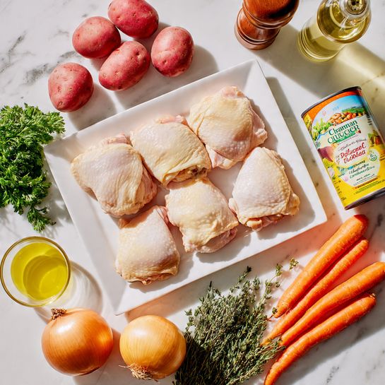 The image shows a white rectangular plate with six raw chicken thighs with pale skin, positioned in two rows of three at the center. Surrounding the plate are fresh ingredients on a white marbled surface: to the top left, a bunch of red potatoes with smooth skins and a few garlic cloves; next to them, green parsley and sprigs of rosemary and thyme with textured leaves; on the right side, a group of six big orange carrots with a smooth texture and a large yellow onion with a papery skin; to the right of the chicken plate, a can of chicken broth with a colorful label and a yellow container of corn starch. Above the plate, there is a clear glass pepper grinder, and to the far right, a green bottle of white wine. A small glass bowl of yellow cooking oil is placed at the lower left corner. All items are neatly arranged with natural lighting, showing detailed textures and vibrant colors photo taken with an iphone --ar 4:5 --v 7
