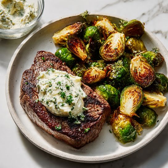 A white plate on a white marbled surface holds a serving of roasted Brussels sprouts and sliced steak. The Brussels sprouts are cut in halves and quarters, showing a mix of bright green and golden-brown crisp edges, scattered on the left side of the plate. On the right side are four thick slices of medium-rare steak, showcasing a brown crust with a pinkish center; the top steak slice is topped with a dollop of creamy herbed butter with visible green flecks. In the upper left corner of the image, a small glass bowl contains more of the creamy herbed butter. The photo taken with an iphone --ar 4:5 --v 7