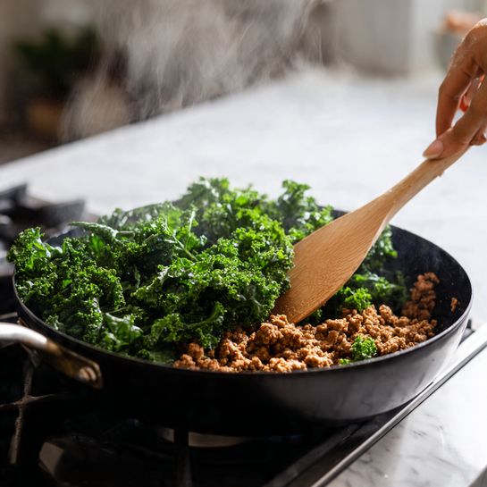 A black frying pan is filled with two visible layers of food on a stove with a white marbled surface. The bottom layer is made of cooked ground meat, shown in a rich brown color with a crumbly texture. On top, a large amount of fresh kale leaves are added, their dark green color contrasting the meat, with steam rising gently from the pan. A wooden spoon held by a woman's hand is stirring the kale, creating movement and focus on the ingredients. The background is softly blurred to keep attention on the pan and food. Photo taken with an iphone --ar 4:5 --v 7