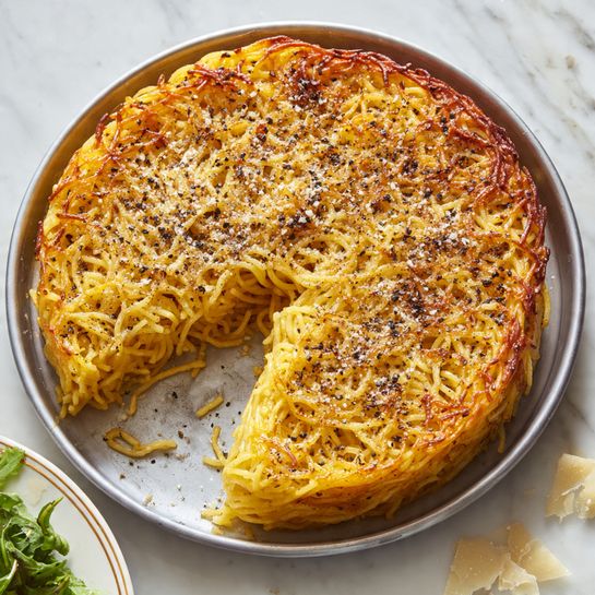 A round baked dish with one layer of spaghetti that is mixed into a creamy light yellow base. The top has some strands of spaghetti that are crisp and golden brown, with black pepper sprinkled evenly across the surface. The edges are slightly browned and the texture shows a combination of smooth creamy parts and crispy pasta loops. The dish is in a round metal pan, placed on a white marbled surface. Photo taken with an iphone --ar 4:5 --v 7