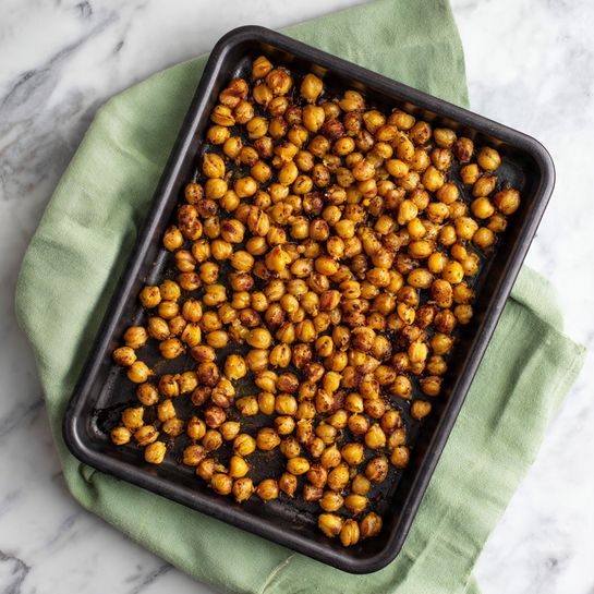 The image shows a black square baking tray filled with a single layer of golden brown roasted chickpeas, spread evenly across the tray. The chickpeas have a slightly crispy texture with some darker spots indicating even roasting. The tray is resting on a light green cloth on top of a white marbled surface. Photo taken with an iphone --ar 4:5 --v 7