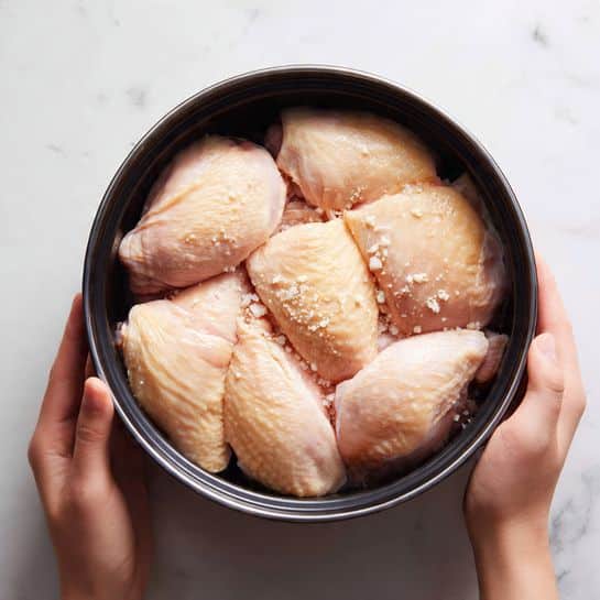 A round black pot filled with several pieces of raw chicken arranged closely together, showing their smooth, pale pink skin with a slight shine. The chicken pieces are layered so that they almost cover the bottom of the pot in one even layer, with some small white fat parts visible between them. The pot is placed on a white marbled surface, and a woman's hand is gently holding the edge of the pot from one side. photo taken with an iphone --ar 4:5 --v 7