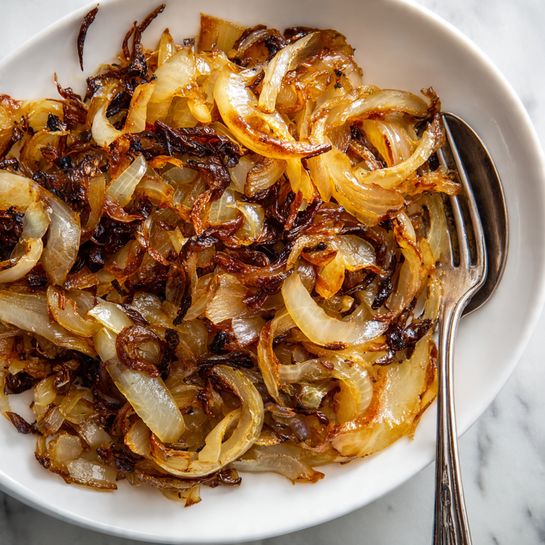 The image shows a close-up of cooked onions with a mix of golden-brown and dark brown colors, some pieces look soft and shiny while others are crispy and slightly charred. The onions are thinly sliced and layered unevenly, creating a texture that looks tender but also crunchy in spots. A silver fork is partly visible on the right side, resting among the cooked onions. The background is a white marbled texture. Photo taken with an iphone --ar 4:5 --v 7