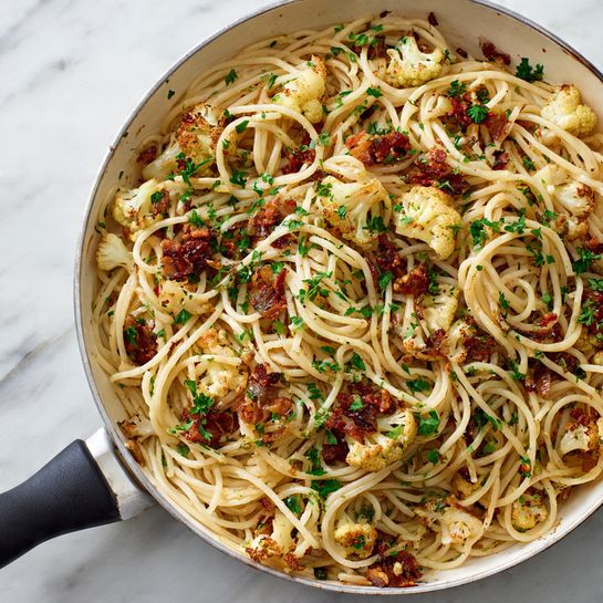 A white pan filled with spaghetti noodles mixed with small pieces of browned cauliflower and bits of crispy, dark brown bacon. Bright green chopped parsley is sprinkled evenly on top, adding fresh color against the light noodles and cauliflower. The spaghetti looks lightly sauced with an oily sheen, and the pan handle is black. The scene is set on a white marbled surface. photo taken with an iphone --ar 4:5 --v 7