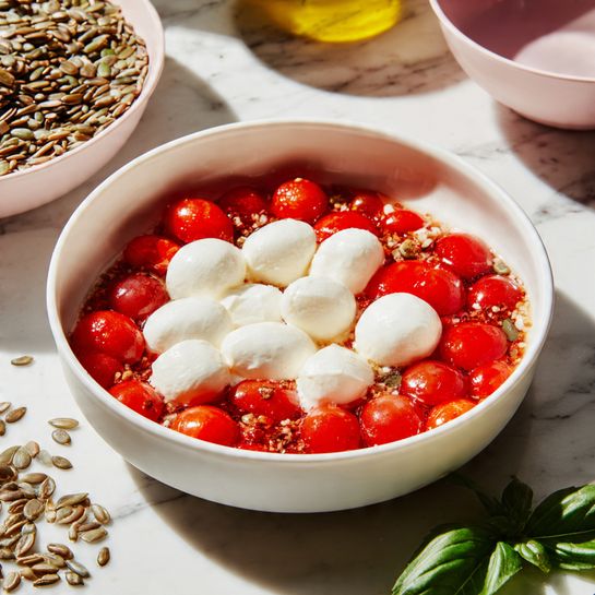 The image shows a white bowl filled with two layers: the bottom layer is made of small, bright red cherry tomato halves with juicy, shiny textures, and on top there is a layer of smooth, white mozzarella balls spread evenly. The bowl is placed on a white marbled surface with scattered green basil leaves nearby and a blurred pink bowl with sunflower seeds in the background. The photo has warm natural light highlighting the fresh ingredients. Photo taken with an iphone --ar 4:5 --v 7