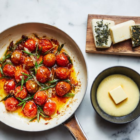 The image shows two parts side by side on a white marbled surface. On the left, a white pan holds cooked small tomatoes, slightly browned and soft, arranged in a single layer with some green herbs and sliced green onions sprinkled on top for color. The tomatoes are surrounded by little burnt bits and orange-red sauce stains on the pan's surface. On the right, a small black pan contains a creamy, pale yellow polenta mixture with a cube of melting butter clearly visible in the center. Behind the pan is a small white wooden board with two small blocks of cheese, one plain and one with dark green herbs. Photo taken with an iphone --ar 4:5 --v 7