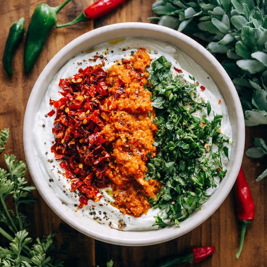 A white round bowl shows a dish with a thick white creamy base spread evenly inside. On top, there are three separate piles: one of bright red finely chopped peppers on the left, one of orange-red chunky paste in the middle, and one of fresh green chopped herbs on the right. The colors are bright and fresh, sprinkled lightly with black pepper. The bowl rests on a wooden surface with some green plants and whole chilies around. photo taken with an iphone --ar 4:5 --v 7