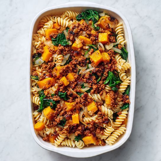 The image shows a white rectangular dish filled with three main layers. The bottom layer is a bed of cooked spiral pasta, light beige in color with a smooth texture. On top of the pasta is a mix of sautéed yellow-orange butternut squash cubes and wilted green spinach leaves, with small bits of translucent cooked onion scattered throughout. The top layer is a generous heap of browned, crumbly ground meat with a rich reddish-brown color, spread unevenly across the vegetable layer. The dish sits on a white marbled surface. photo taken with an iphone --ar 4:5 --v 7