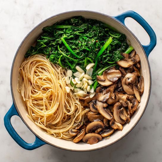 A white round pot with blue handles is shown from the top on a white marbled surface. Inside the pot, three main layers are visible: bright green leafy vegetables on one side, light brown thin pasta in the middle, and sliced brown mushrooms on the other side. Small pieces of white garlic are sprinkled on top of the pasta. The ingredients look fresh and lightly cooked, sitting in a clear broth at the bottom. photo taken with an iphone --ar 4:5 --v 7