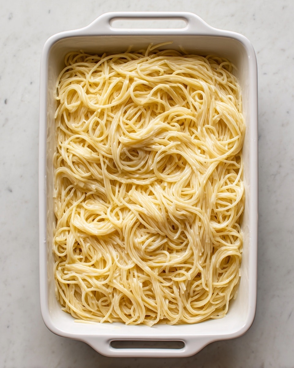 A white rectangular baking dish filled with one layer of creamy spaghetti noodles, the noodles are pale yellow and coated evenly with a smooth, light cream sauce, creating a glossy look. The dish sits on a white marbled textured surface. photo taken with an iphone --ar 4:5 --v 7