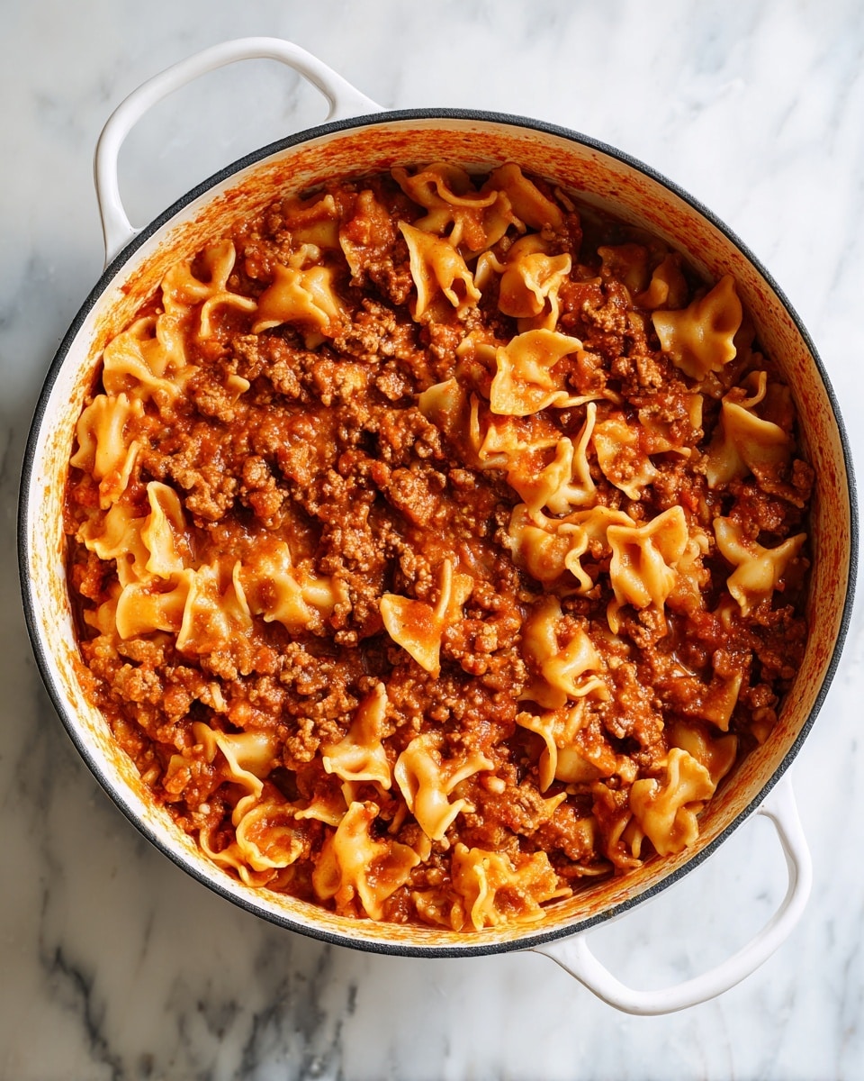 A white plate holds a dish with two main layers: large, curved pasta pieces on the bottom, light orange with a ribbed texture, mixed with a thick layer of chunky meat sauce on top. The sauce is rich in a red-orange color with visible small carrots and ground meat pieces, creating a thick, hearty texture that coats the pasta well. The plate is on a white marbled surface with a rustic wooden background above. Photo taken with an iphone --ar 4:5 --v 7
