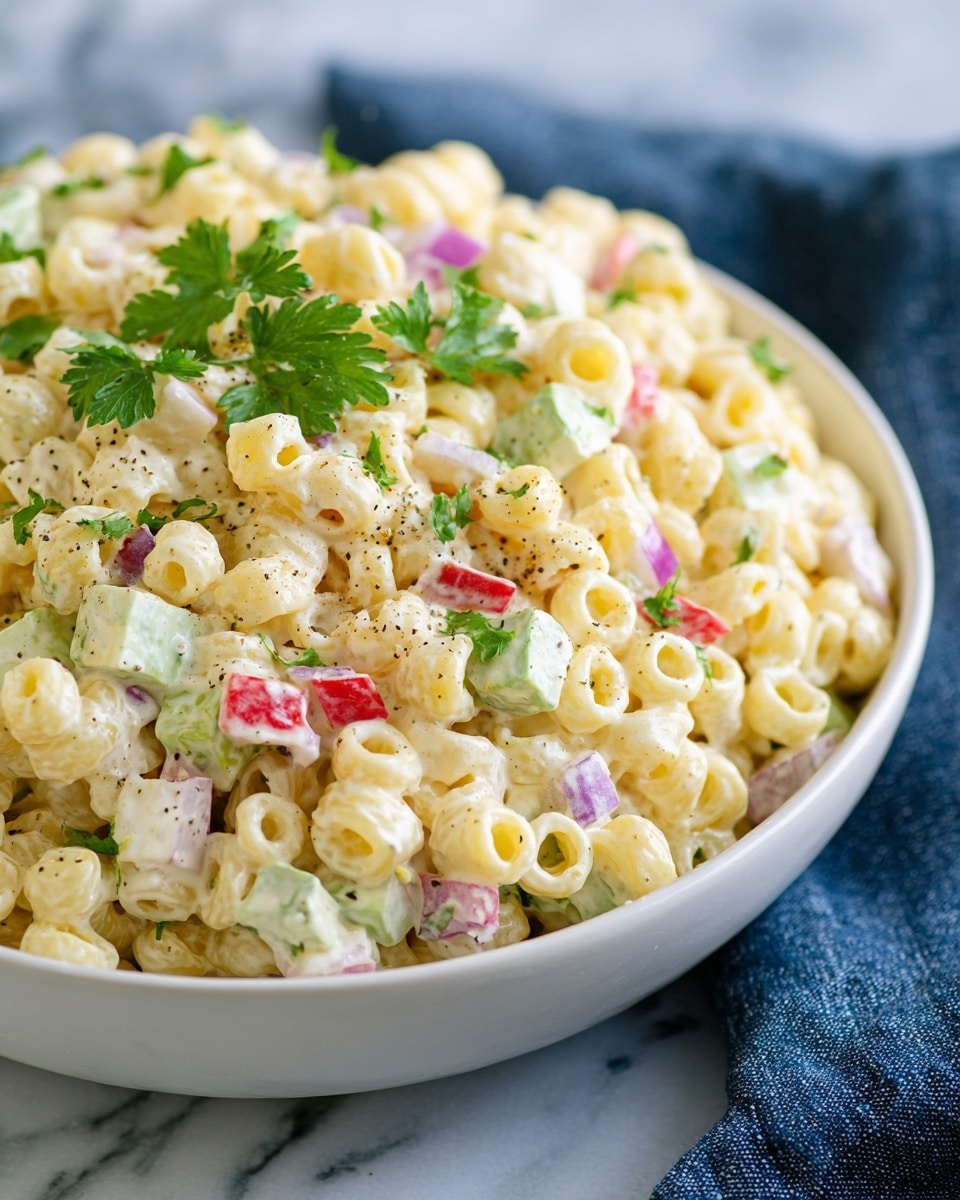 A close-up view of a white bowl filled with a creamy pasta salad. The salad has small, round pasta rings as the main layer, which are pale yellow and soft in texture. Mixed evenly throughout the pasta are small pieces of red bell pepper, green celery, chopped boiled egg whites with yellow yolks, and bits of purple onion, adding splashes of color. The whole salad is coated with a smooth, light cream dressing and topped with fresh, green parsley leaves and a light sprinkle of black pepper. The bowl is set on a white marbled surface with a dark blue cloth in the background. photo taken with an iphone --ar 4:5 --v 7