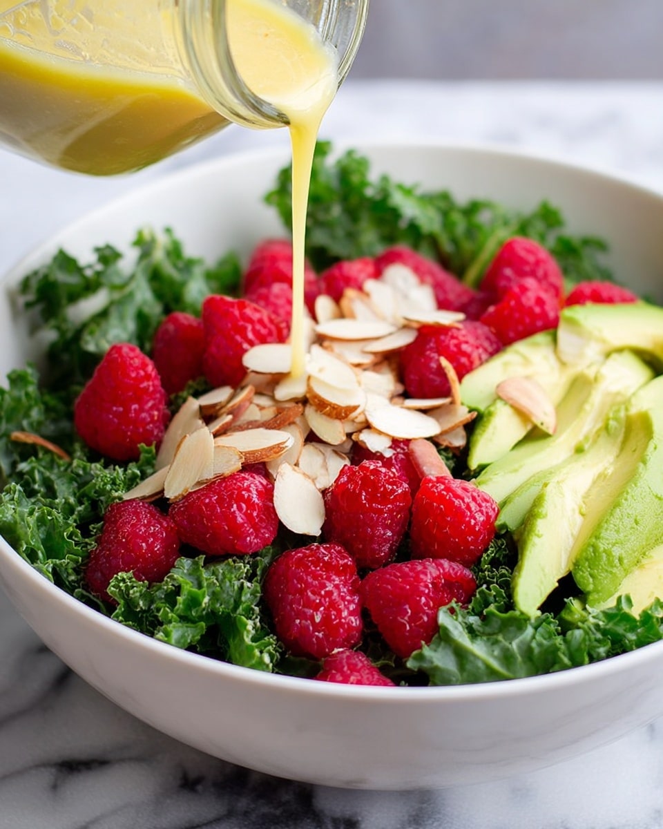The image shows a fresh salad in a white bowl on a white marbled surface. The bottom layer is dark green kale leaves with a rough, curly texture. On top of the kale, there are bright red raspberries that look soft and juicy, along with slices of light green avocado that are smooth and creamy. Scattered over the salad are thin, slightly toasted almond slices with a beige color and light brown edges. A light yellow dressing is being poured over the salad from a glass jar, adding a smooth, glossy layer starting at the center. Photo taken with an iphone --ar 4:5 --v 7