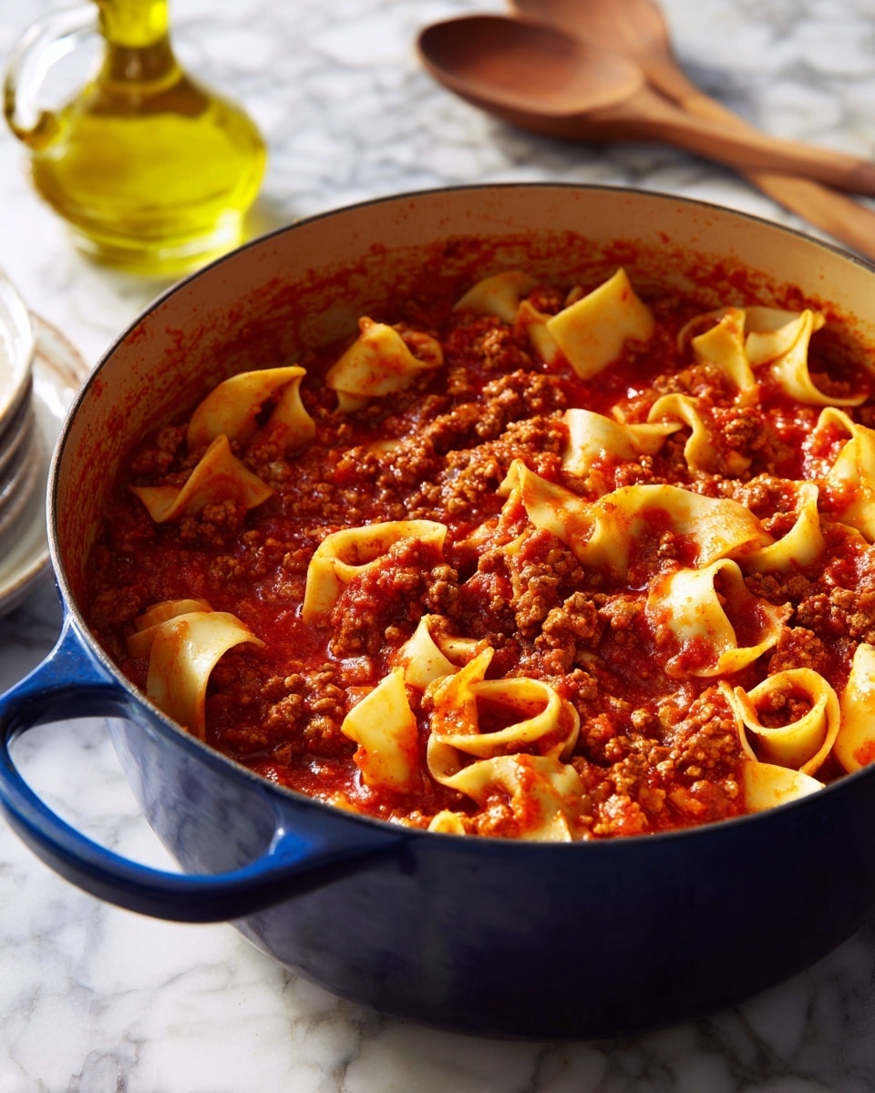 A deep blue pot filled with a thick red tomato sauce holding small brown chunks of ground meat and wide, flat pasta pieces with wavy edges, all simmering gently on a stove. The sauce has a smooth and slightly chunky texture, coating the pasta and meat well. The pot sits on a white marbled kitchen surface, with a clear glass bottle containing yellow oil and a wooden spoon nearby. The scene feels warm and inviting, showing a hearty pasta meal in the making. photo taken with an iphone --ar 4:5 --v 7