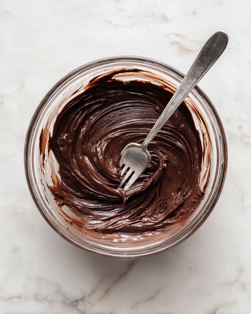 A clear glass bowl sits on a white marbled surface, filled with a single layer of dark brown, thick, and slightly grainy mixture that looks smooth and glossy. A silver fork rests inside the bowl with its tines partly covered in the mixture, positioned diagonally from the bottom right toward the center of the bowl. The mixture shows swirled patterns made by the fork’s motion, giving the surface a textured appearance with some lighter brown spots around the edges photo taken with an iphone --ar 4:5 --v 7