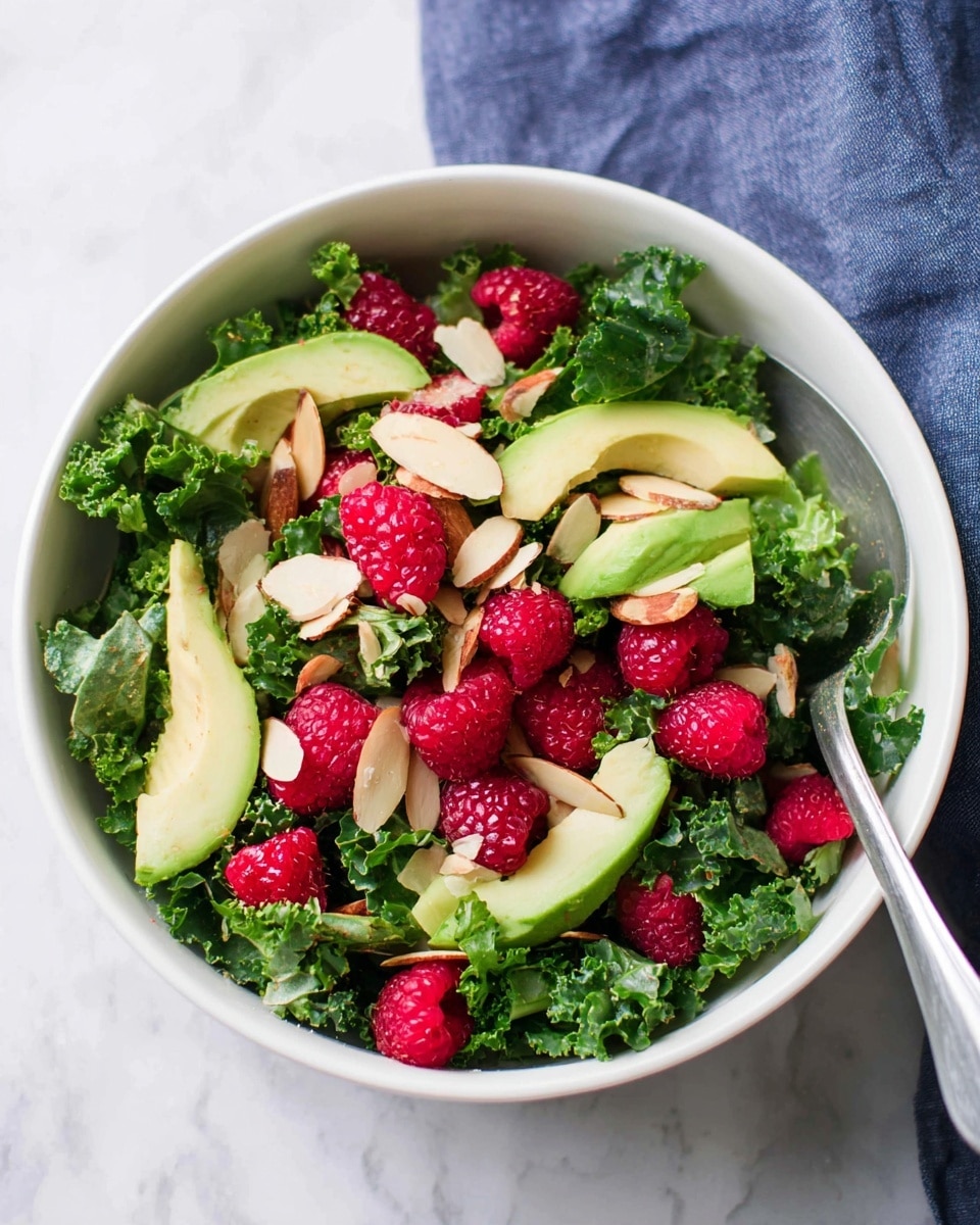 A fresh salad served in a white bowl with three visible layers: the bottom layer is dark green curly kale leaves with a textured, leafy surface; the middle layer has light green avocado slices with smooth, creamy texture scattered evenly; the top layer is bright red raspberries with a bumpy surface and thin, light brown almond slices sprinkled across. The bowl is placed on a white marbled surface. photo taken with an iphone --ar 4:5 --v 7