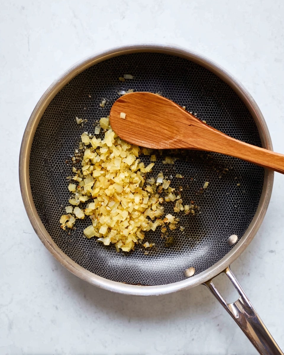 A stainless steel frying pan with a honeycomb-textured black cooking surface holds a single layer of finely chopped onions being cooked to a golden-brown color. A long wooden spoon lies inside the pan, resting diagonally from the upper right edge towards the center, its smooth light brown surface contrasting with the darker pan base. The pan handle extends to the right, shiny and metallic, sitting on a white marbled surface. photo taken with an iphone --ar 4:5 --v 7