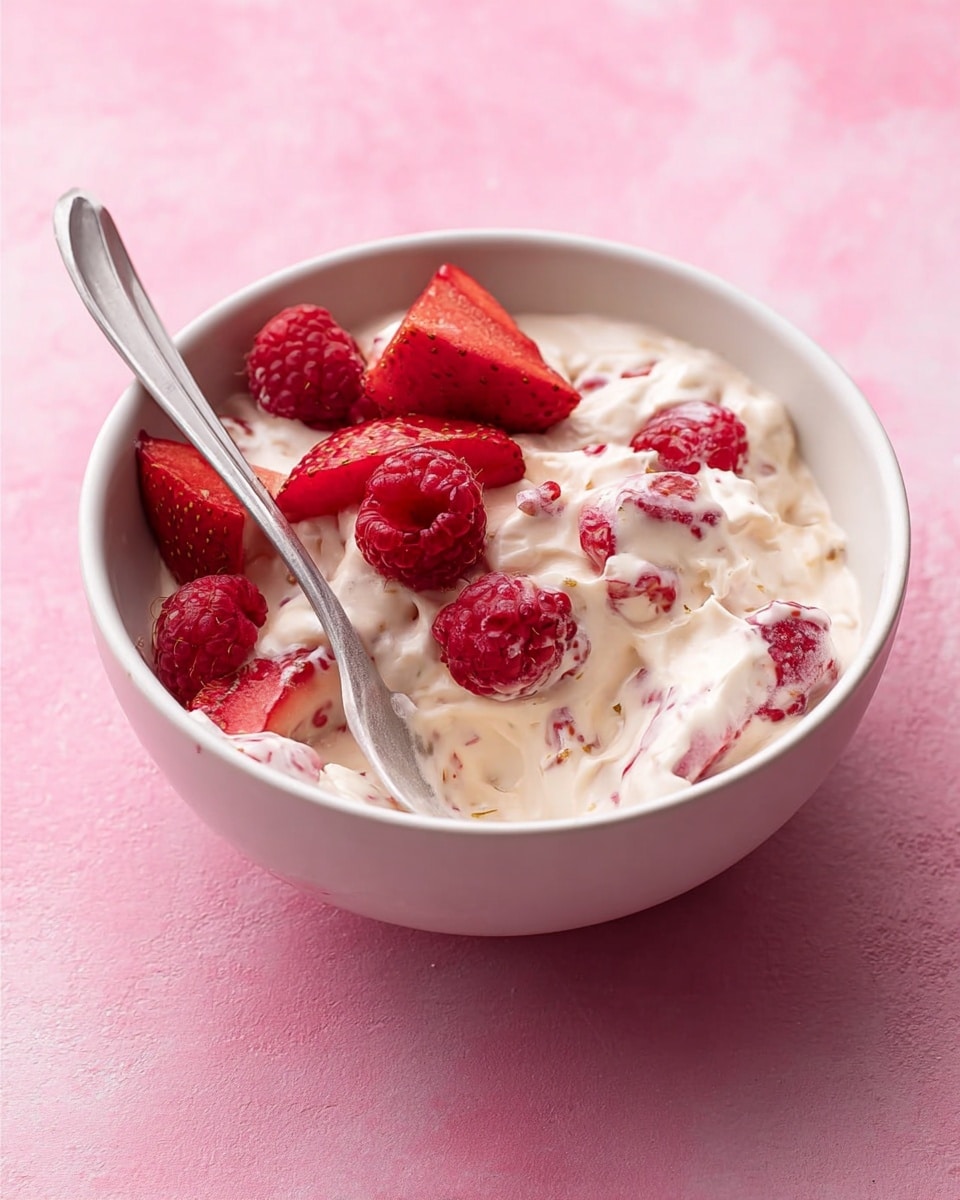 The image shows a clear glass bowl filled with a creamy mixture that is mostly white with visible pieces of red fruit mixed throughout. The texture of the creamy mixture looks thick and smooth with soft swirls, while the red fruit pieces appear small and uneven, adding spots of color. The bowl sits on a white marbled surface with a soft pink background, and the mixture looks fresh and lightly whipped, giving a airy impression. Photo taken with an iphone --ar 4:5 --v 7