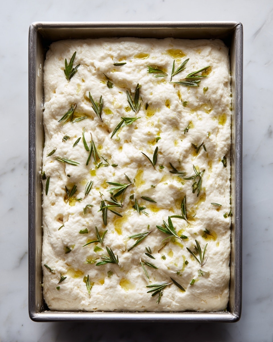 The image shows a rectangular metal baking pan filled with risen dough that has a soft and airy texture with many bubbles and uneven surfaces. Scattered across the top are fresh green rosemary leaves adding a touch of color contrast. The dough appears lightly brushed with olive oil, making parts of its white surface shine slightly. The pan is placed on a white marbled textured surface, and the overall look is rustic and fresh, ready for baking. photo taken with an iphone --ar 4:5 --v 7