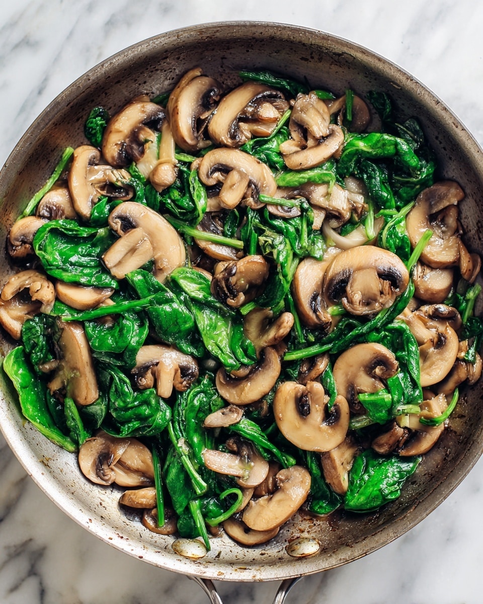 The image shows a pan filled with cooked mushrooms and spinach. The mushrooms are light brown and have a soft, moist texture. They are mixed evenly with bright green spinach leaves that look tender and slightly wilted. The vegetables sit directly in the pan, showing a simple cooked dish with no separate layers. The background is a white marbled surface. photo taken with an iphone --ar 4:5 --v 7